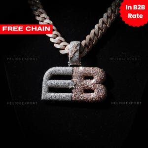 Op de afbeelding: Een met diamanten bezette hangerketting met de letters "EB" in zilver en roségoud. De hanger hangt aan een dikke, bijpassende ketting. De afbeelding bevat ook de tekst "FREE CHAIN" en "In B2B Rate".