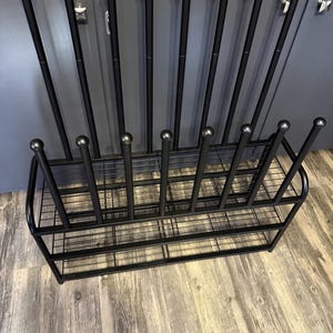 May include: Black metal boot rack with a rectangular base and vertical supports. The rack has a grid-like design for the base and a series of upright rods with ball-shaped tops. The rack is designed to hold multiple pairs of boots.