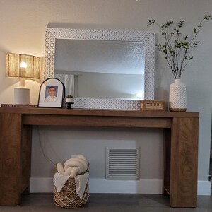 May include: A wooden console table with a large mirror and decorative items. The mirror has a white patterned frame. A lamp, a framed photo, and a vase with greenery sit on the table. A woven basket with a blanket is below.