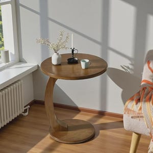May include: A round wooden side table with a curved C-shaped base. The tabletop holds a white ceramic vase with small white flowers, a black metal candle holder with a white taper candle, and a small light blue ceramic mug with a handle.