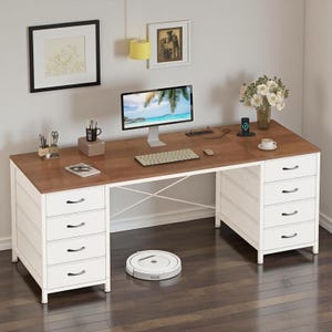 May include: A large wooden desk with a brown top and white frame, featuring two side cabinets with four drawers each. The desk holds a computer monitor, keyboard, mouse, and office supplies. A white robotic vacuum sits on the floor underneath.