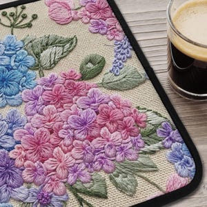 May include: Embroidered floral placemat featuring a hydrangea design in shades of pink, purple, and blue. The placemat has a black border and is set on a light-colored, textured surface. A glass of coffee is visible in the background.