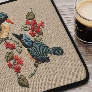 以下が含まれることがあります： 赤い花が咲く枝に止まっている2羽のカラフルな鳥が刺繍されたベージュのプレースマット。鳥は青、オレンジ、黒のディテールがあります。黒い縁取り。背景にはコーヒーが入ったグラスがあります。