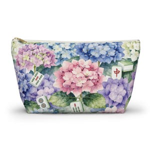 Könnte beinhalten: Eine Reißverschlusstasche mit einem Aquarelldruck aus rosa, blauen und violetten Hortensienblüten, durchsetzt mit verstreuten Mahjong-Steinen. Die Steine zeigen verschiedene Symbole und das rote chinesische Schriftzeichen für Mitte vor Blumen.