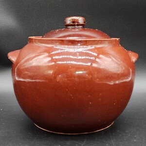 Op de afbeelding: Een ronde, donkerbruine keramische pot met een bijpassend deksel en twee kleine handvatten aan de zijkanten. Het oppervlak heeft een glanzende, gladde afwerking met subtiele variaties in het bruine glazuur. De pot heeft een brede opening.