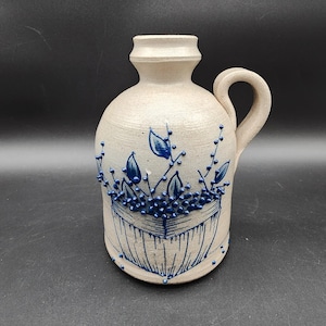Pode incluir: Um jarro de cerâmica bege com uma única asa e um gargalo estreito. O corpo apresenta um design em relevo pintado a azul de um cesto cheio de ramos com folhas e pequenas bagas, com uma textura marcada sobre a superfície mate do objeto.