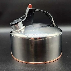 May include: Stainless steel stovetop kettle with a black handle and spout, and a copper-colored base. The kettle has a red knob on the lid and a black spout cover. The polished metal reflects the surrounding environment.