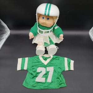 Könnte beinhalten: Eine Cabbage Patch Kids Puppe mit blondem Wollhaar, blauen Augen und roten Wangen, bekleidet mit einem weißen Football-Helm mit grünen Streifen, einem grünen Langarmshirt und einem weißen Rock. Ein grünes Football-Trikot mit der Nummer 27 liegt bei.