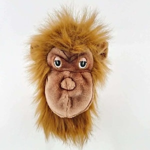 May include: A soft toy monkey head, featuring brown fur and a detailed face. The monkey has a light brown muzzle, dark eyes, and a slightly open mouth. The shaggy fur frames the face, giving it a playful and whimsical look.