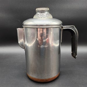 Puede incluir: Una cafetera percoladora de acero inoxidable con acabado plateado pulido, asa negra resistente al calor, base de tono cobre y un pomo de cristal transparente en la tapa. El cuerpo cilíndrico presenta un pico lateral para verter.