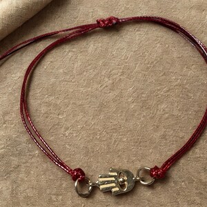 Op de afbeelding: Een bordeauxrode armband met een Hamsa hand bedel. De armband is gemaakt van dun, gevlochten koord en heeft een zilverkleurige sluiting. De Hamsa hand bedel is gedetailleerd met lijnen en een kleine lus.