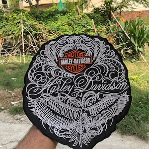 Peut inclure: Un écusson brodé noir de forme circulaire avec un motif en filigrane blanc, un aigle central et le logo orange et blanc Harley-Davidson Motor Cycles. Le texte Harley-Davidson est brodé en écriture blanche au centre de l'écusson.