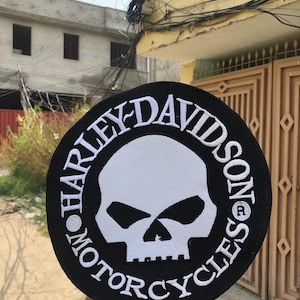 Puede incluir: Un parche bordado circular en blanco y negro que presenta un gráfico de calavera blanca en el centro. El texto HARLEY-DAVIDSON MOTORCYCLES está bordado en letras mayúsculas blancas alrededor del borde negro que rodea la calavera.