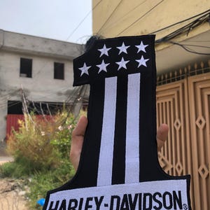 May include: A black and white embroidered patch in the shape of the number one. The top section features seven white stars, while the vertical bar contains two white stripes. The base displays the text HARLEY-DAVIDSON in white capital letters.