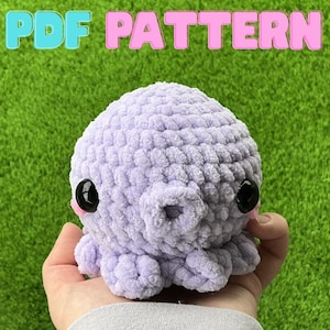 May include: A handmade, lavender crochet octopus with black safety eyes and a round, open mouth. The text "PDF PATTERN" is displayed in the top center of the image. The octopus is held against a green background.