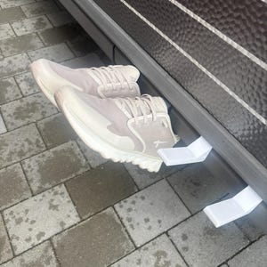 Könnte beinhalten: Ein Paar hellbeige Sportschuhe mit weißen Sohlen und Schnürsenkeln. Die Schuhe befinden sich in der Nähe einer dunkelbraunen Oberfläche mit weißen Streifen. Sie ruhen auf weißen Kunststoffstützen.