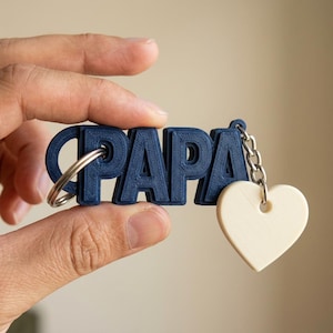 May include: A navy blue keychain spelling "PAPA" with a silver keyring and a cream-colored heart charm. The keychain is held in a hand against a neutral background. The heart charm is attached to the keychain with a silver chain.