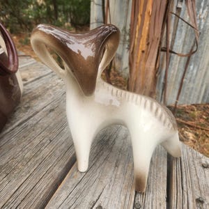 Può includere: Statuetta di ariete in ceramica con testa e corna marroni lucide e corpo color crema. La statuetta ha un design stilizzato ed è posizionata su una superficie di legno. Una borsa marrone e crema è parzialmente visibile sullo sfondo.