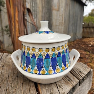 Può includere: Una zuppiera in ceramica bianca con coperchio, con un motivo di pesci blu, gialli e bianchi. La zuppiera ha due manici e poggia su una superficie di legno. Il coperchio ha un piccolo disegno blu e giallo.