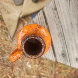 Op de afbeelding: Bovenaanzicht van een oranje keramische kruik met een handvat. De kruik heeft een donkere opening en een getextureerd oppervlak. De kruik staat tegen een achtergrond van verweerd hout en droog gebladerte.
