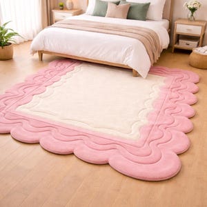 May include: A pink and cream-colored rug with a scalloped edge sits on a hardwood floor in front of a bed. The rug has a wavy border and a rectangular center. The bedroom has a neutral color palette.