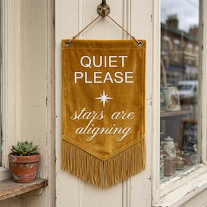 Puede incluir: Bandera de color mostaza con las palabras "QUIET PLEASE" y "stars are aligning" en escritura blanca. Un gráfico de estrella está entre las frases. La bandera tiene flecos en la parte inferior y cuelga de un gancho de metal.