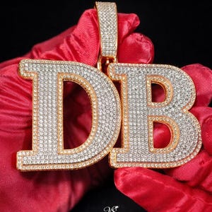 Könnte beinhalten: Ein goldfarbener Anhänger mit den Buchstaben "DB", besetzt mit Diamanten. Die Buchstaben sind goldfarben umrandet und mit kleinen, funkelnden Steinen gefüllt. Der Anhänger wird vor rotem Hintergrund präsentiert.