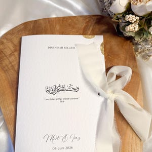 Puede incluir: Una invitación de boda blanca con detalles dorados y una cinta crema. La invitación presenta caligrafía en idioma extranjero y los nombres "Mert & Jaz" con la fecha "06. Juni 2026". Las palabras "DINI NIKAH BELGESI" están en la parte superior.