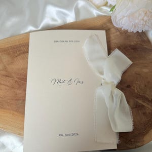 Puede incluir: Un programa de boda color crema con el texto "DINI NIKAH BELGESI", los nombres "Mert & Iraz" y la fecha "06. Juni 2026". Está atado con una cinta color crema. El fondo incluye una superficie de madera y flores blancas.