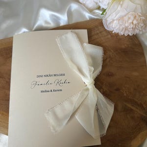 Pode incluir: Certificado de casamento cor marfim amarrado com uma fita creme. O certificado tem caligrafia elegante com os nomes Melisa & Kerem. O fundo inclui flores brancas e uma superfície de madeira.