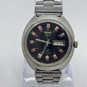 Op de afbeelding: Een vintage HMT Rajat polshorloge met een roestvrijstalen band en kast. De wijzerplaat is zwart met rode en witte markeringen en de dag wordt op 3 uur weergegeven. Het horloge is gemarkeerd met "AUTOMATIC 21 JEWELS".