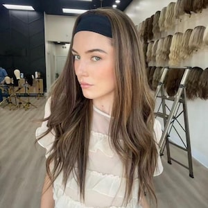May include: A woman with long, wavy brown hair with blonde highlights, wearing a black headband and a white top. The background shows a wall of wigs and a metal ladder.