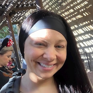 May include: A woman with long, dark hair and a black headband smiles at the camera. She wears a leopard print top and a gray backpack. The background includes a wooden structure and other people.