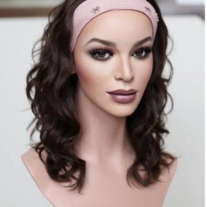 May include: A light pink fabric headband featuring small silver-toned bow embellishments, worn on a mannequin head with shoulder-length, wavy dark brown hair. The headband is positioned across the forehead, securing the dark brown hair back.