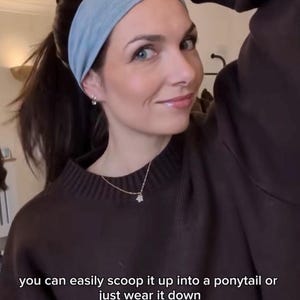 May include: A woman wearing a light blue headband with a dark brown sweater. The headband is made of a soft, stretchy material. The text on the image says, "you can easily scoop it up into a ponytail or just wear it down."