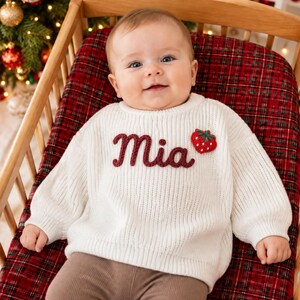 May include: A white long-sleeve ribbed knit sweater featuring the name Mia embroidered in dark red yarn across the chest, accompanied by a small red strawberry appliqué with green leaves positioned to the right of the name.