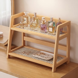 Puede incluir: Estantería de madera de dos niveles con acabado claro, que contiene varios frascos de perfume y una pequeña bandeja de joyas. La estantería tiene un diseño perforado y está sobre una superficie de madera.