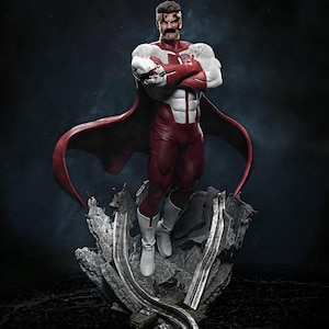 Op de afbeelding: Gedetailleerd beeldje van een superheld in een rood-wit kostuum, staand op een grijze, rotsachtige basis. Het figuur heeft een rode cape en witte laarzen. De superheld heeft een snor en een gezichtsverwonding.