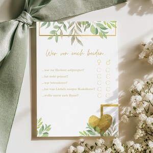 May include: A wedding game card with the text "Wer von euch beiden..." in gold script. The card features a gold frame, watercolor leaves, and a gold heart. A sage green ribbon and white flowers are in the background.