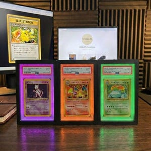 May include: A black display case holding three illuminated Pok&eacute;mon trading cards. The cards feature Mewtwo, Charizard, and Venusaur, each in a graded holder with purple, orange, and green backlights.