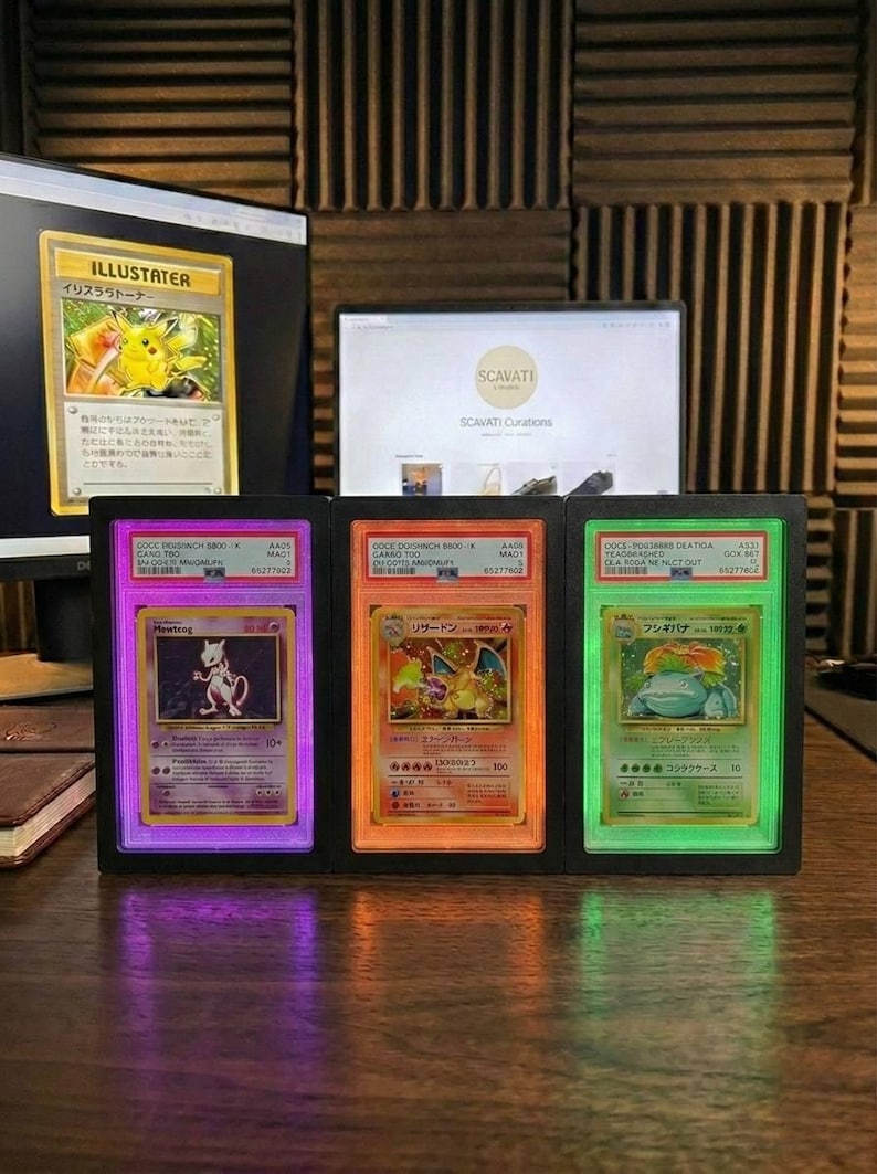 May include: A black display case holding three illuminated Pok&eacute;mon trading cards. The cards feature Mewtwo, Charizard, and Venusaur, each in a graded holder with purple, orange, and green backlights.