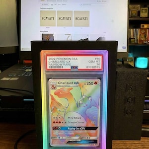 May include: A graded 2022 Pok&eacute;mon Charizard GX Rainbow Rare card in a black display case with a colourful LED border. The card features a vibrant illustration of Charizard. The display case is in front of a computer screen.