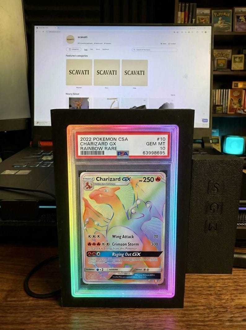 May include: A graded 2022 Pok&eacute;mon Charizard GX Rainbow Rare card in a black display case with a colourful LED border. The card features a vibrant illustration of Charizard. The display case is in front of a computer screen.