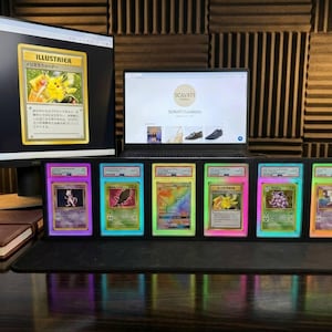 May include: Graded Pok&eacute;mon trading cards in protective cases with neon borders are displayed. A computer monitor and laptop are in the background, showing a Pok&eacute;mon card image and a website. The cards are arranged on a desk.
