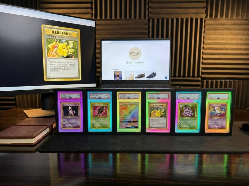 May include: Graded Pok&eacute;mon trading cards in protective cases with neon borders are displayed. A computer monitor and laptop are in the background, showing a Pok&eacute;mon card image and a website. The cards are arranged on a desk.