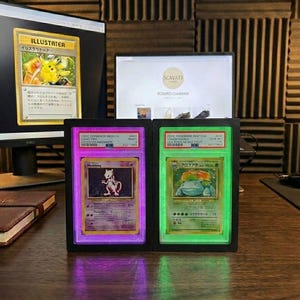 May include: Two illuminated display cases showcasing collectible Pok&eacute;mon trading cards. One case glows purple, displaying a Mewtwo card, and the other glows green, displaying a Venusaur card. The cards are encased in protective holders.
