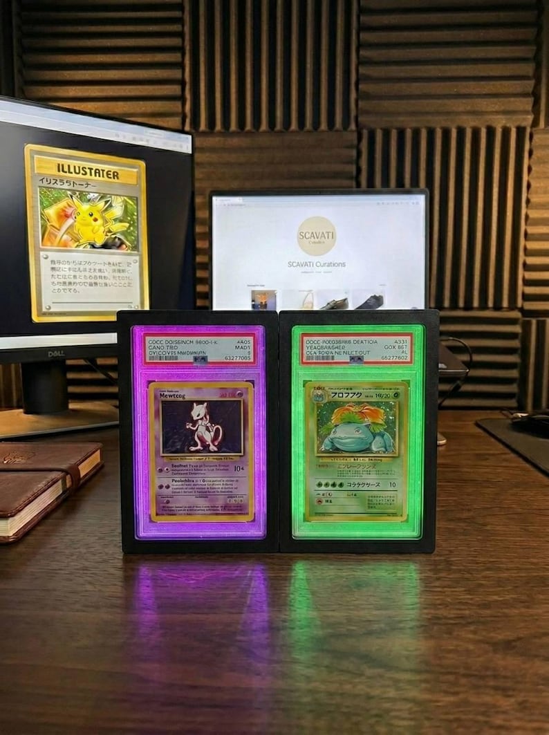 May include: Two illuminated display cases showcasing collectible Pok&eacute;mon trading cards. One case glows purple, displaying a Mewtwo card, and the other glows green, displaying a Venusaur card. The cards are encased in protective holders.