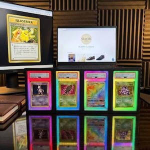 May include: A collection of graded Pok&eacute;mon trading cards, showcased with vibrant LED lighting. The display features various cards, including a Pikachu Illustrator card on a computer screen. A laptop displays the Scavati Curations website.