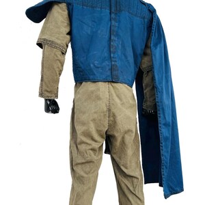 Back view of a Gar Saxon Mandalorian costume on a model, highlighting the long blue cape and the rear construction of the flak vest. Shows the full-length drape of the fabric and the tan flight suit pants, set against a professional white background.