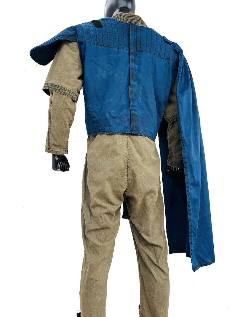 Back view of a Gar Saxon Mandalorian costume on a model, highlighting the long blue cape and the rear construction of the flak vest. Shows the full-length drape of the fabric and the tan flight suit pants, set against a professional white background.